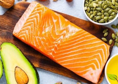 Composição harmoniosa de alimentos ricos em gorduras boas: salmão fresco, abacate cortado ao meio, variedade de oleaginosas (nozes, amêndoas, pistache), sementes de abóbora em tigela de madeira e azeite de oliva em pequeno recipiente. Dispostos sobre superfície clara, representando as principais fontes de ácidos graxos insaturados essenciais para saúde cardiovascular e cerebral.