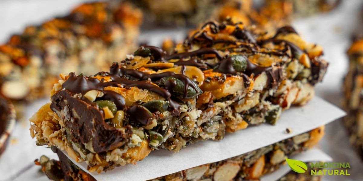 Barras energéticas caseiras densamente recheadas com uma variedade impressionante de nozes inteiras e picadas, sementes nutritivas e frutas secas naturais, criando uma textura crocante e satisfatória. A imagem destaca a riqueza nutricional incomparável das barras feitas com ingredientes à base de plantas, onde cada pedaço visível de amêndoa, castanha, semente de girassol e cranberry contribui com seu perfil único de vitaminas, minerais e antioxidantes.