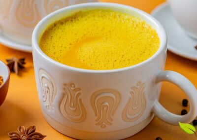 Xícara de vidro com chá de cúrcuma Golden Milk decorado com canela em pó, acompanhado de paus de canela sobre superfície clara. Esta versão aromática combina as propriedades anti-inflamatórias da cúrcuma com o calor da canela, criando uma bebida reconfortante e terapêutica. Ideal para fortalecer o sistema imunológico e melhorar a digestão.