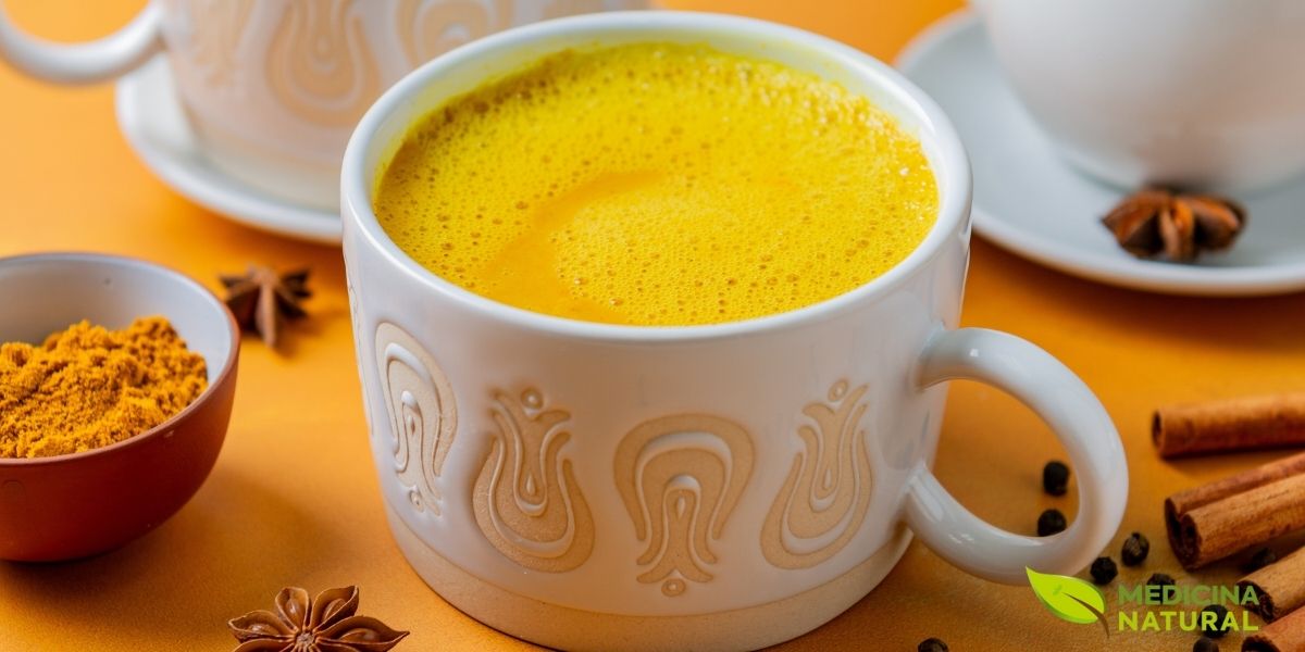 Xícara de vidro com chá de cúrcuma Golden Milk decorado com canela em pó, acompanhado de paus de canela sobre superfície clara. Esta versão aromática combina as propriedades anti-inflamatórias da cúrcuma com o calor da canela, criando uma bebida reconfortante e terapêutica. Ideal para fortalecer o sistema imunológico e melhorar a digestão.
