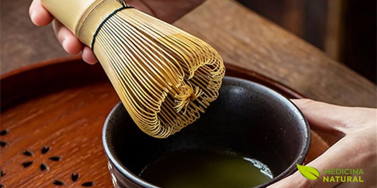 Chasen de bambu natural em uso sobre tigela preta de matcha, demonstrando a técnica correta de movimento em W para incorporar ar e criar a espuma perfeita. O batedor deve ser movido rapidamente mas sem tocar o fundo da tigela, criando um padrão que mistura o pó de chá com a água de forma uniforme. Após o uso, o chasen deve ser enxaguado com água fria e colocado em um suporte (kusenaoshi) para manter sua forma circular. Com cuidado adequado, um chasen de qualidade pode durar meses de uso regular.