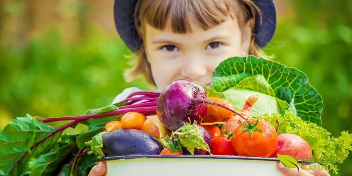 Uma criança sorridente segura orgulhosamente uma cesta repleta de vegetais coloridos, incluindo brócolis, repolho roxo, tomates e pimentões. Esta imagem representa perfeitamente o conceito da "dieta do arco-íris", onde cada cor de vegetal oferece um conjunto único de vitaminas, minerais e fitonutrientes essenciais para o crescimento saudável. Incentivar as crianças a explorar essa variedade de cores é uma estratégia lúdica e eficaz para garantir uma nutrição completa e equilibrada.
