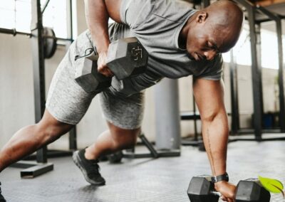 Atleta executando exercício composto com halteres em academia moderna, demonstrando a versatilidade dos treinos com pesos livres para o desenvolvimento muscular completo. Os exercícios com halteres permitem uma maior amplitude de movimento e recrutamento de fibras musculares estabilizadoras, gerando um estímulo mais completo para a hipertrofia. A nutrição pós-treino, incluindo proteínas de rápida absorção e carboidratos, é crucial para otimizar a recuperação e maximizar os ganhos de massa muscular. Fonte: Muscle & Fitness.