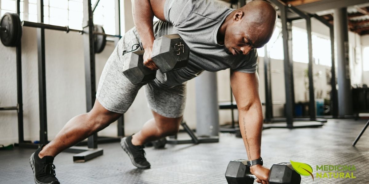 Atleta executando exercício composto com halteres em academia moderna, demonstrando a versatilidade dos treinos com pesos livres para o desenvolvimento muscular completo. Os exercícios com halteres permitem uma maior amplitude de movimento e recrutamento de fibras musculares estabilizadoras, gerando um estímulo mais completo para a hipertrofia. A nutrição pós-treino, incluindo proteínas de rápida absorção e carboidratos, é crucial para otimizar a recuperação e maximizar os ganhos de massa muscular. Fonte: Muscle & Fitness.